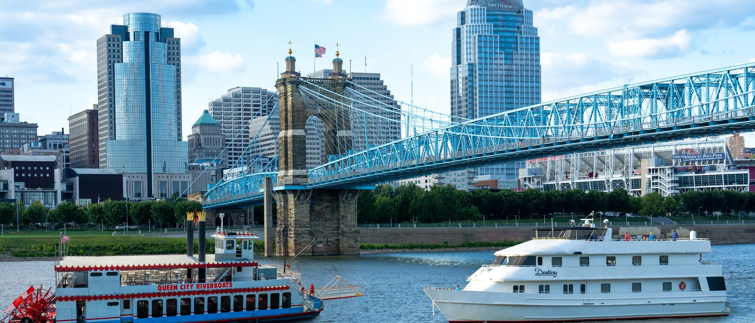 The Spirit of Cincinnati paddlewheeler and the Destiny yacht side by side on the Ohio River under the Roebling Suspension Bridge, the Cincinnati skyline behind them, with the Queen City Riverboats and Destiny Yacht Charters logos in the sky